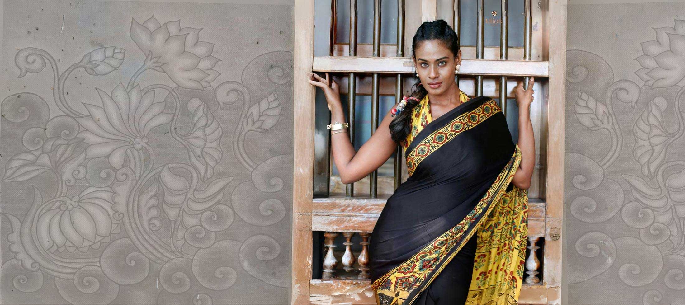 Woman in a black and gold saree standing in front of a decorative wall.